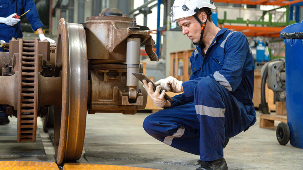 Maintenance worker in blue coveralls and helmet kneels beside large industrial machinery, inspecting a component with a tablet in hand in a factory setting